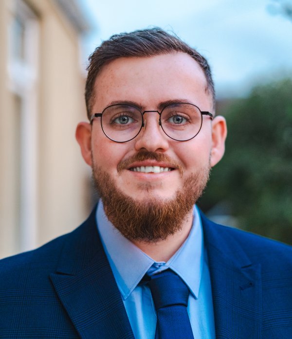 Portrait of a smiling bearded man with glasses, wearing a blue suit and tie, with a blurred building in the background.