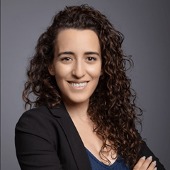 Portrait of a smiling woman with curly hair wearing a white blouse, set against a plain light background.