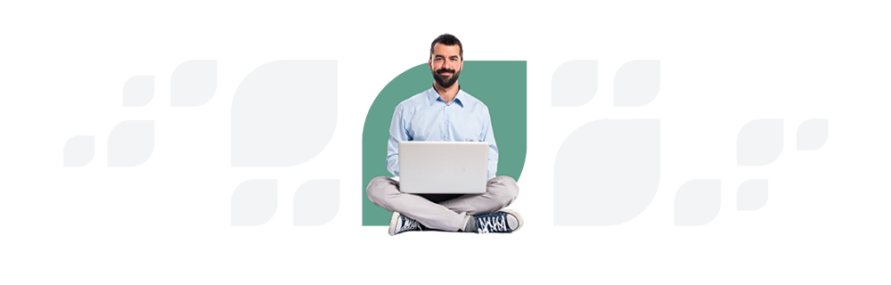 Man with a beard sitting cross-legged, holding a laptop, smiling at the camera, dressed in a blue shirt and gray pants.