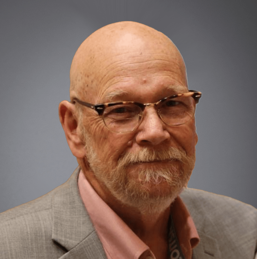 Elderly man with a full beard and glasses, wearing a pink blazer, smiling gently at the camera in an indoor setting.