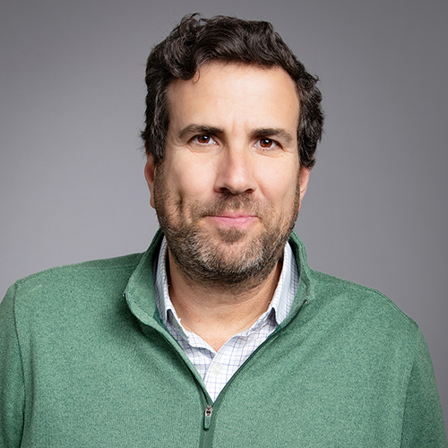 A middle-aged man with stubble, wearing a green sweater over a checkered shirt, poses against a gray background, giving a subtle smile.