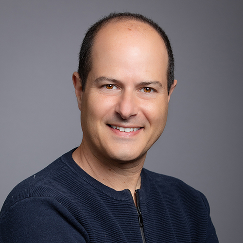 Middle-aged man smiling at the camera, wearing a navy sweater, against a grey background.