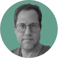 A middle-aged man with glasses and stubble, wearing a gray t-shirt, set against a teal background.